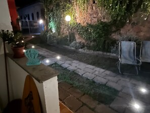 A paved pathway leading to a stone wall with greenery, a potted plant, and a metal chair.