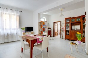 Un comedor con una mesa de madera, sillas blancas, un televisor y una zona de cocina con armarios y un refrigerador.