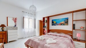 Ein Schlafzimmer mit Bett, Nachttisch, Kommode, Stuhl, Lampe, einem Gemälde und einem Fenster mit Vorhängen.