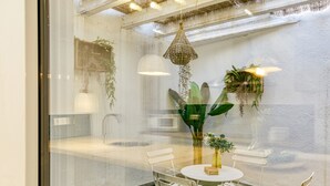A room with a glass wall, a round table with chairs, hanging plants, and a hanging light fixture. - Apartment 'Ca La Maca' with Wi-Fi and Air Conditioning (Llançà)