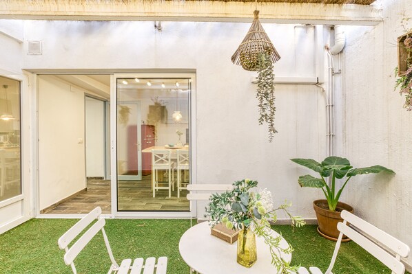 Une terrasse avec un revêtement de gazon synthétique, une table blanche, un banc, un luminaire suspendu et une plante en pot.