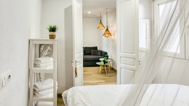 A bedroom with a bed, a shelf with towels, a small table with a plant, and a view into another room with a sofa and hanging lights. - Apartment 'Ca La Maca Mediterrani' with Mountain View, Wi-Fi and Air Conditioning (Llançà)