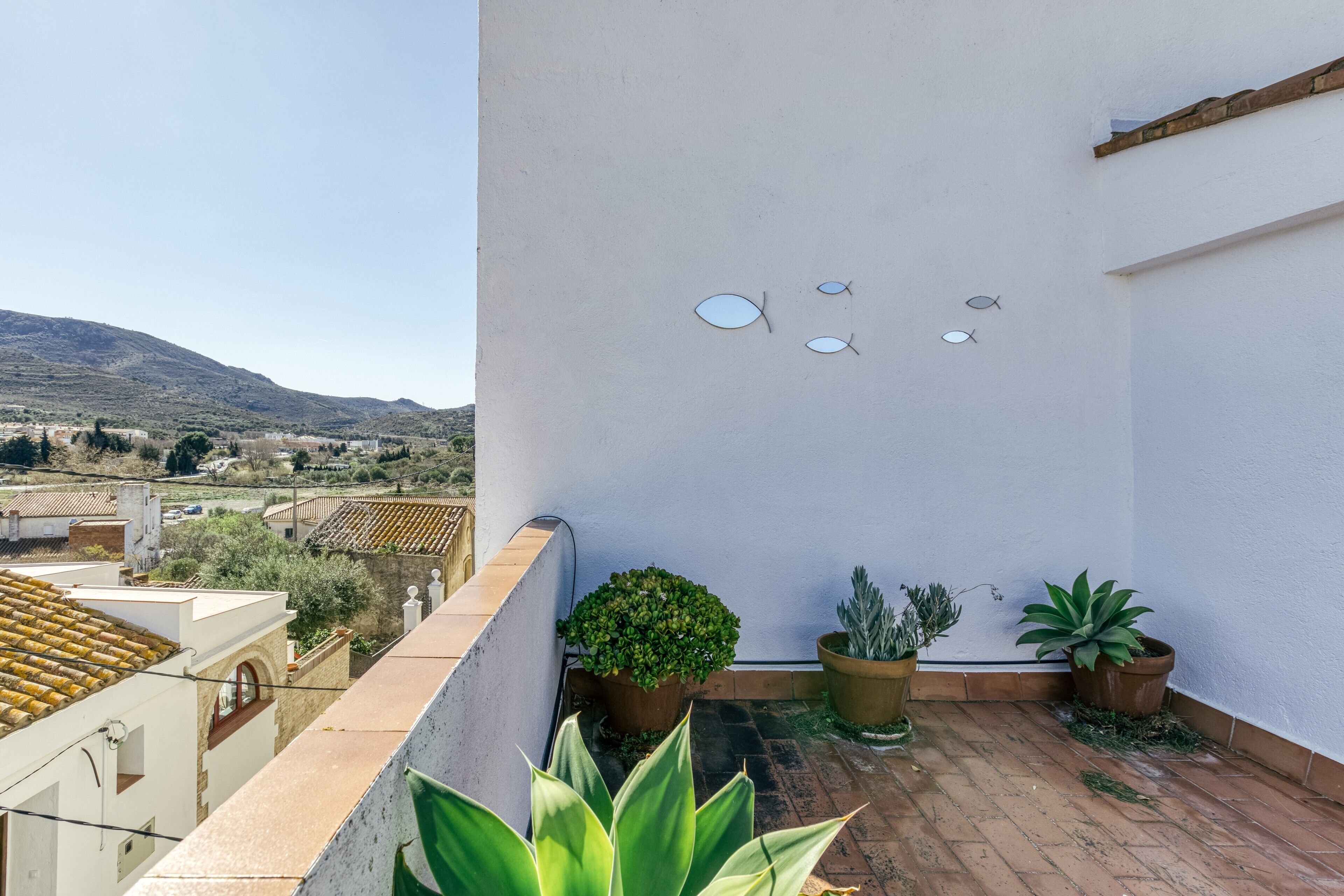Terraza en la azotea con plantas en macetas, una pared blanca con adornos de peces y vistas a colinas distantes.