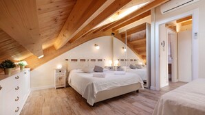 Ein Schlafzimmer mit schrägen Wänden, Holzbalken, einem Bett mit weißer Bettwäsche, Nachttischen mit Lampen und einer kleinen Pflanze.
