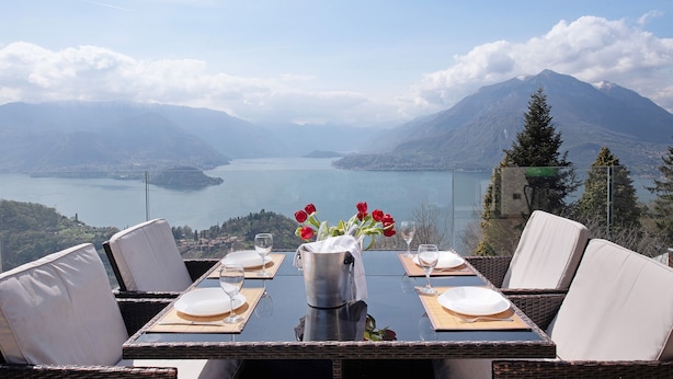 A dining table with white chairs, a vase with red flowers, and a view of a lake and mountains. - Holiday Home "Relax e Felicità" Lake View, Private Jacuzzi, Shared Heated Indoor Pool (Perledo)