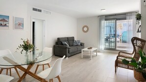 A modern living room with a glass dining table, white chairs, a grey sofa, and a wicker chair. - Family Apartment 'Coral' with Private Pool, Wi-Fi and Air Conditioning (Conil de la Frontera)
