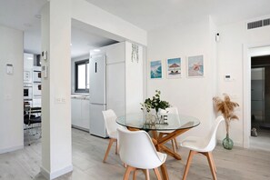 Un espace repas moderne avec une table en verre, des chaises blanches et une cuisine en arrière-plan.
