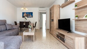 Un salon moderne comprenant un canapé, un téléviseur, un meuble de rangement en bois, une table à manger, des chaises et un tableau représentant un bâtiment.