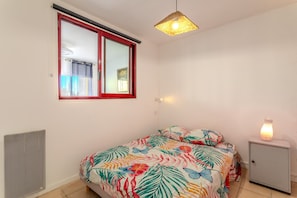 Un dormitorio con cama, ventana de marco rojo, calentador en la pared, mesita de noche con lámpara y un armario pequeño.