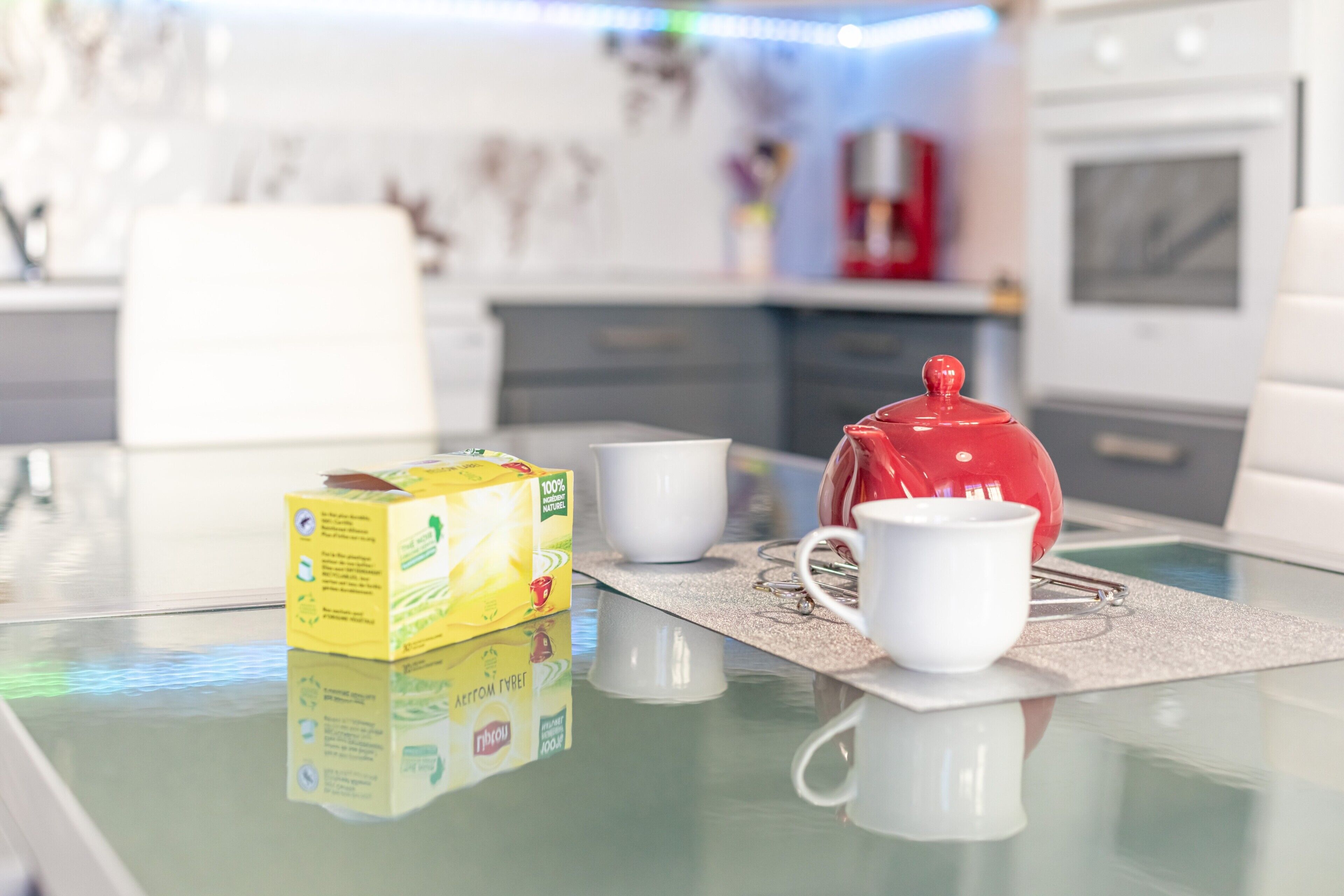 A modern kitchen with a glass table, a red teapot, a white cup, and a box of tea.