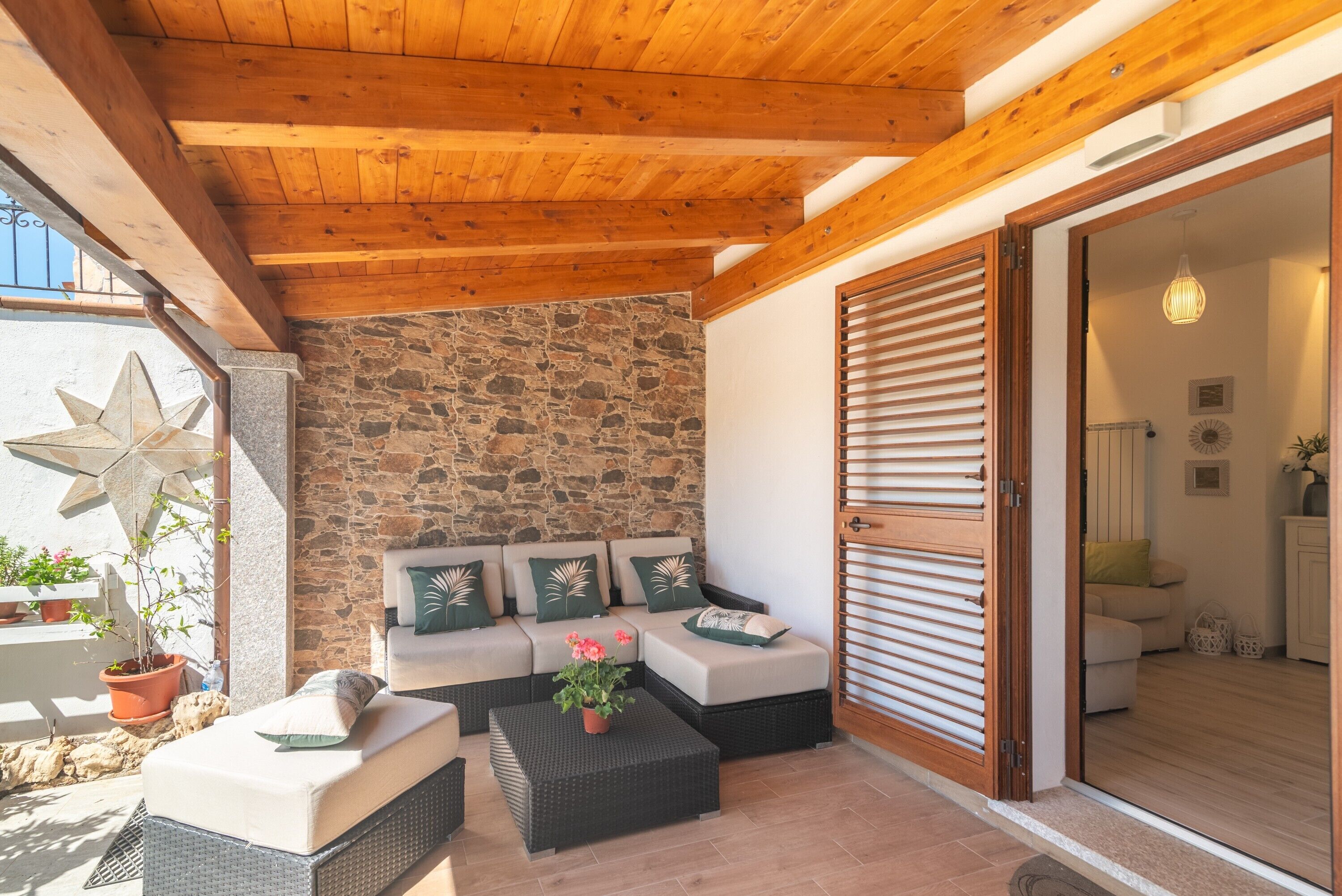 Un patio cubierto con pared de piedra, vigas de madera en el techo y un conjunto de sofás, además de una planta en maceta.