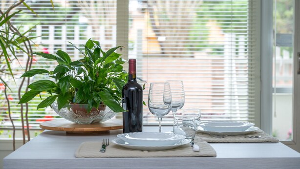 Mesa lista para comer, con platos, copas de vino y una botella, una planta en maceta y una ventana con persianas en el fondo.