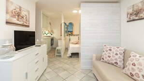 Una habitación funcional con cama, sofá, televisión, zona de cocina y obras de arte en las paredes.