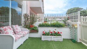 Un balcón con césped artificial, un sofá de mimbre blanco y plantas en macetas.