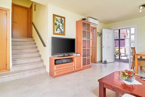 A living room with a TV on a wooden stand, a staircase, a dining table with a vase, and a door leading outside.