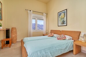 A bedroom with a bed, a window with curtains, a bedside table with a lamp, a wall-mounted painting, and a small wooden stool.