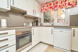 A kitchen with white cabinets, a built-in oven, a dishwasher, and a window with a floral curtain.