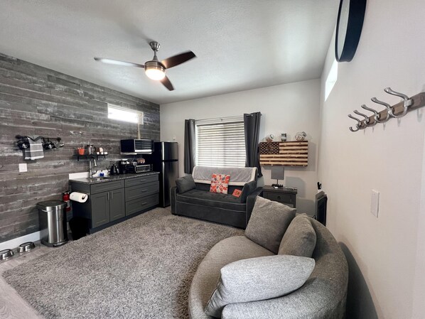 A compact living space with a kitchenette, a ceiling fan, a sofa, and a window with blinds.
