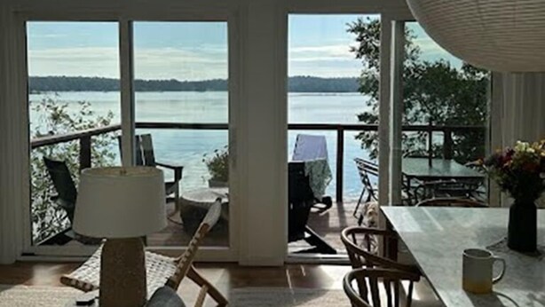 A spacious room with large windows offering a view of a lake and distant trees. There is a dining table with chairs and a lamp. - Coveshead. Dramatic waterfront designer cottage. (Harpswell)