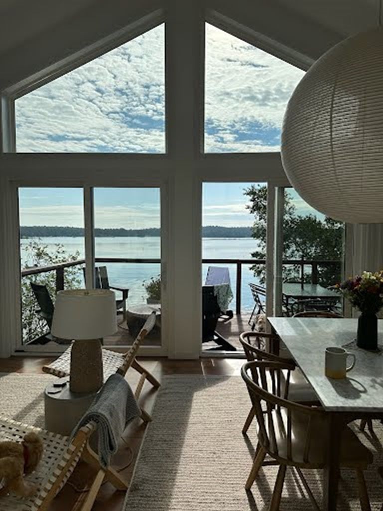 A spacious room with large windows offering a view of a lake and distant trees. There is a dining table with chairs and a lamp.