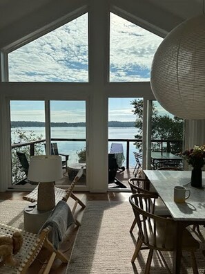 Ein geräumiges Zimmer mit großen Fenstern und Blick auf einen See und ferne Berge. Ein Esstisch mit Stühlen und eine Lampe sind vorhanden.