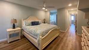 Une chambre à coucher comprenant un lit, des tables de chevet, une commode, un ventilateur de plafond et un couloir menant à une salle de bain.