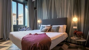 A hotel room with a large bed, a window with curtains, a round side table with a lamp, and a patterned floor. - Sky Suite - My Home in Como (Como)