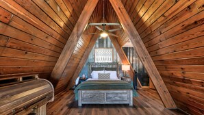 Une chambre mansardée chaleureuse avec un plafond en pente, des murs en bois, un lit recouvert d’une couverture verte, une table de chevet et un ventilateur de plafond.