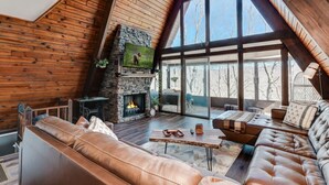 L’intérieur chaleureux d’un chalet avec une cheminée en pierre, des canapés en cuir et de grandes fenêtres donnant sur la forêt environnante.
