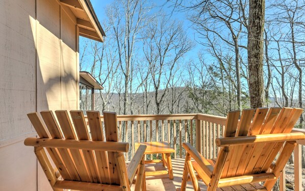 Una terraza de madera con sillas Adirondack y una pequeña mesa, con vista a un área boscosa.
