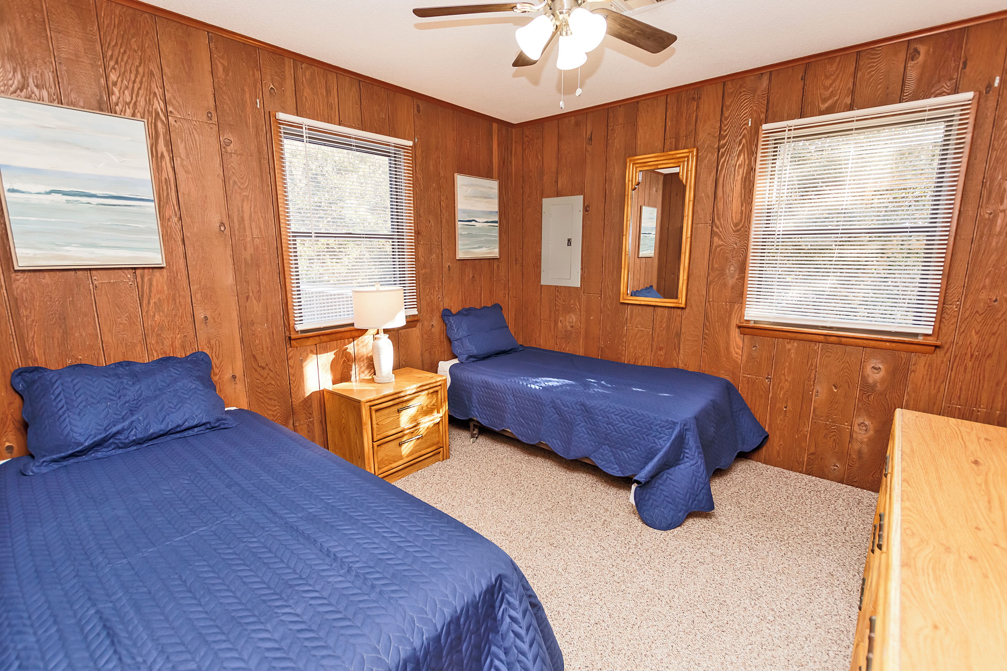 A room with wooden paneling, two beds with blue bedding, a ceiling fan, and a window with blinds.