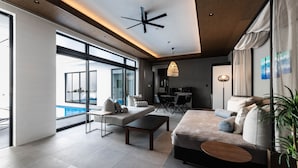 A modern living room with a sofa, coffee table, and dining area. There is a large window with a view of a pool. - CLASS THE FIRST (Miyakojima)