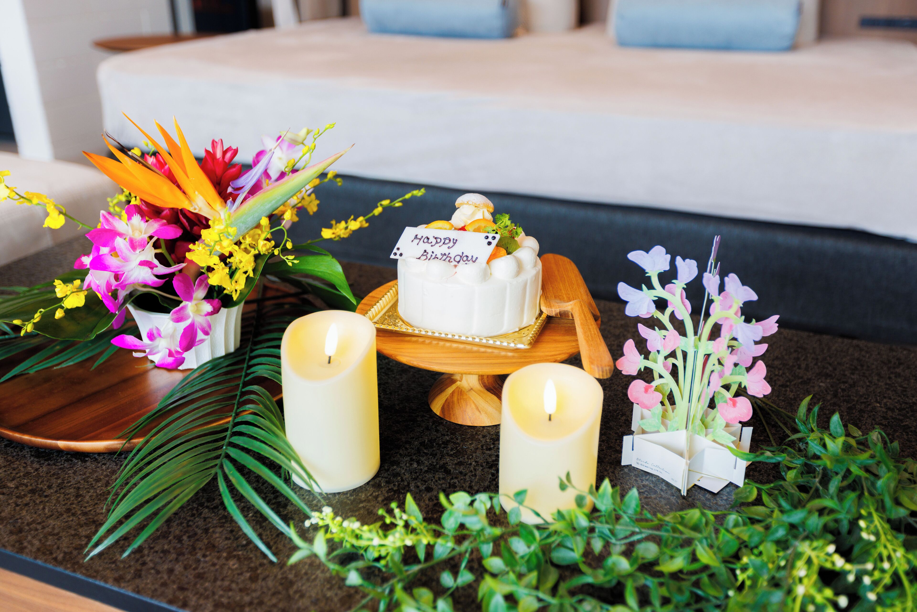 A table with a white cake on a wooden stand, surrounded by candles, flowers, and a 'Happy Birthday' card.