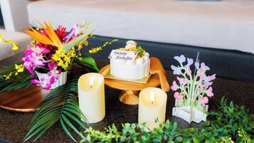 A table with a white cake on a wooden stand, surrounded by candles, flowers, and a 'Happy Birthday' card.