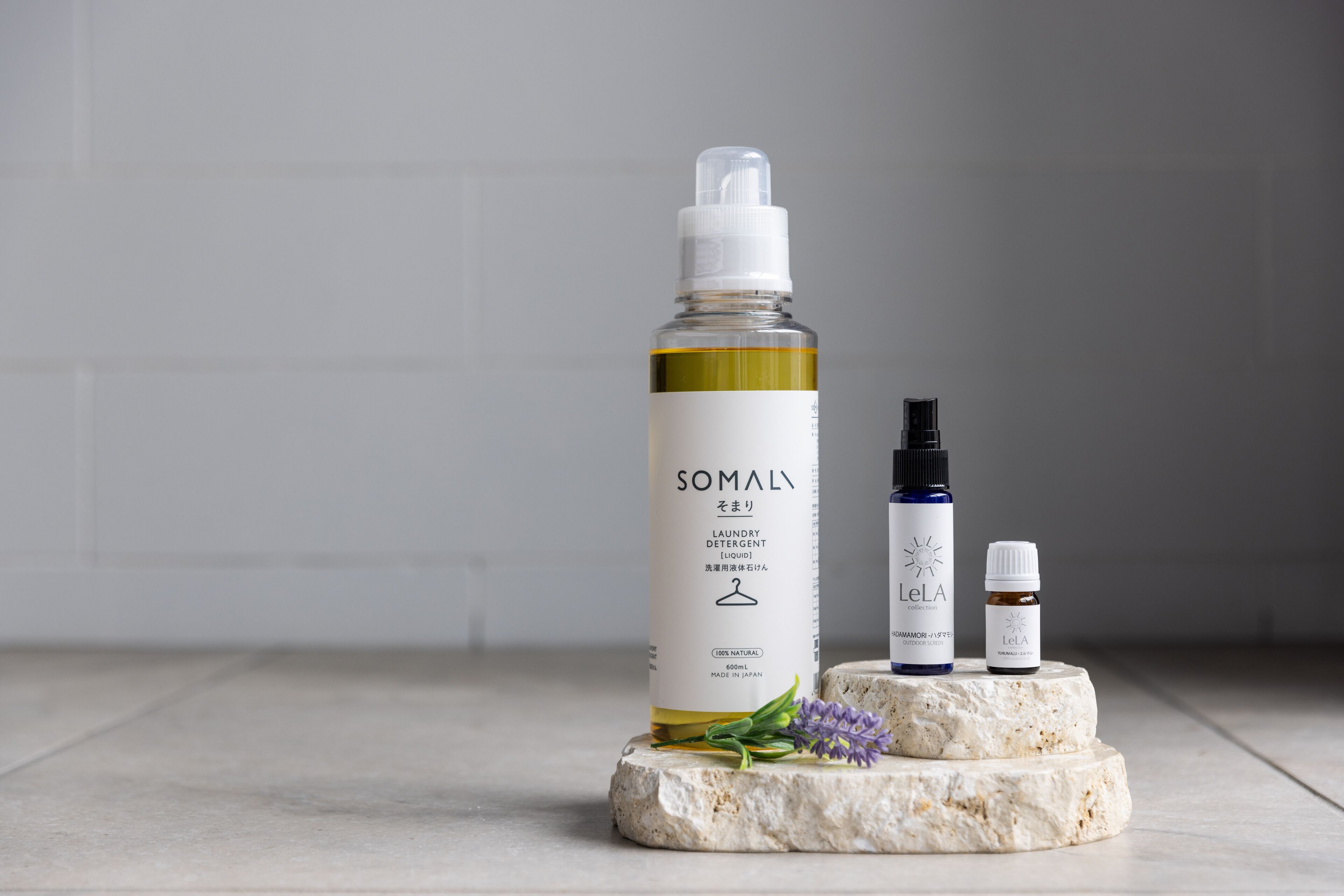 Une bouteille de lessive SOMALY, une bouteille de la collection LeLA, et une petite bouteille posée sur une surface en pierre, le tout sur un fond blanc.