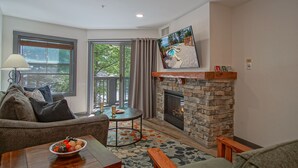 A cozy living room with a stone fireplace, a comfortable sofa, a coffee table with fruit, and a view of the outdoors through a large window. - Rimfire 259 : Walking distance to village,slopes, 1 Br Sleeps 4 (Snowshoe)