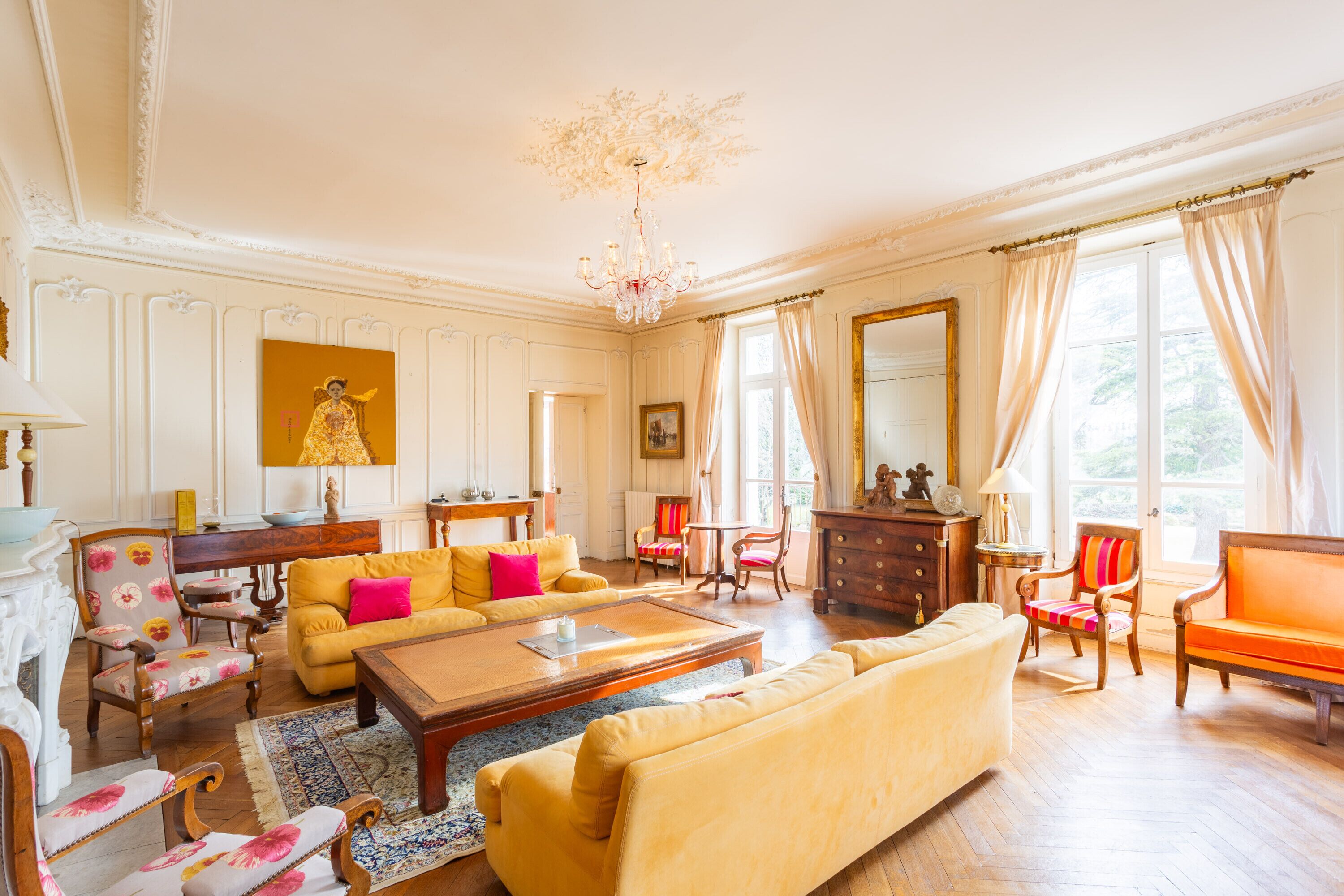 A spacious living room with a yellow sofa, a wooden coffee table, and a chandelier. There are multiple chairs and a fireplace.