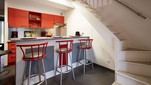 A modern kitchen with red cabinets, a built-in island with bar stools, and a staircase leading to an upper level. - Maison Labennaise - Welkeys (Labenne)