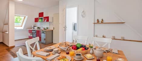 A dining area with a wooden table set for breakfast, a kitchen in the background, and a view of the outdoors through a window.