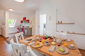 A dining area with a wooden table set for breakfast, a kitchen in the background, and a view of the outdoors through a window.