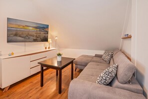 Ein modernes Wohnzimmer mit Holzboden, einem grauen Sofa mit gemusterten Kissen, einem kleinen Tisch und einem Gemälde vom Sonnenuntergang über einem Strand.