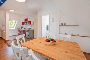 Un comedor con mesa de madera y sillas blancas, una cocina con armarios rojos, y un pequeño balcón con vistas.