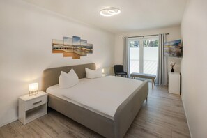 A modern bedroom with a large bed, bedside tables, a chair, a small table with a vase, and artwork on the wall.