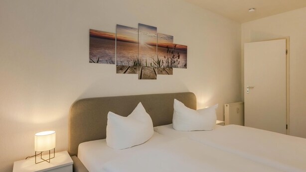 Ein Hotelzimmer mit einem Bett, zwei Kissen, einer Nachttischlampe, einem Nachttisch und einem Wandbild mit Sonnenuntergang über Wasser.