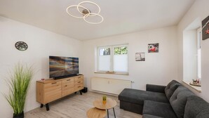 Ein modernes Wohnzimmer mit einem Flachbildfernseher, einem grauen Sofa, einem Holzschrank und einem kleinen Tisch mit einer Pflanze.