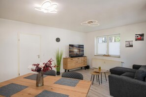 A modern living room with a wooden dining table, grey sofas, a TV on a wooden cabinet, and a window with blinds.