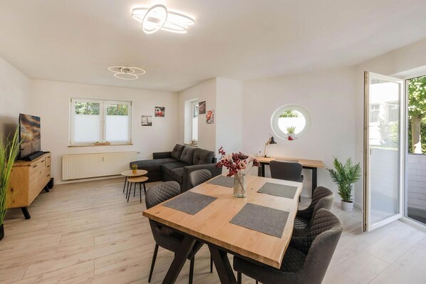 A modern living room with a sofa, a dining table, and a TV. There are windows and a view of greenery outside.