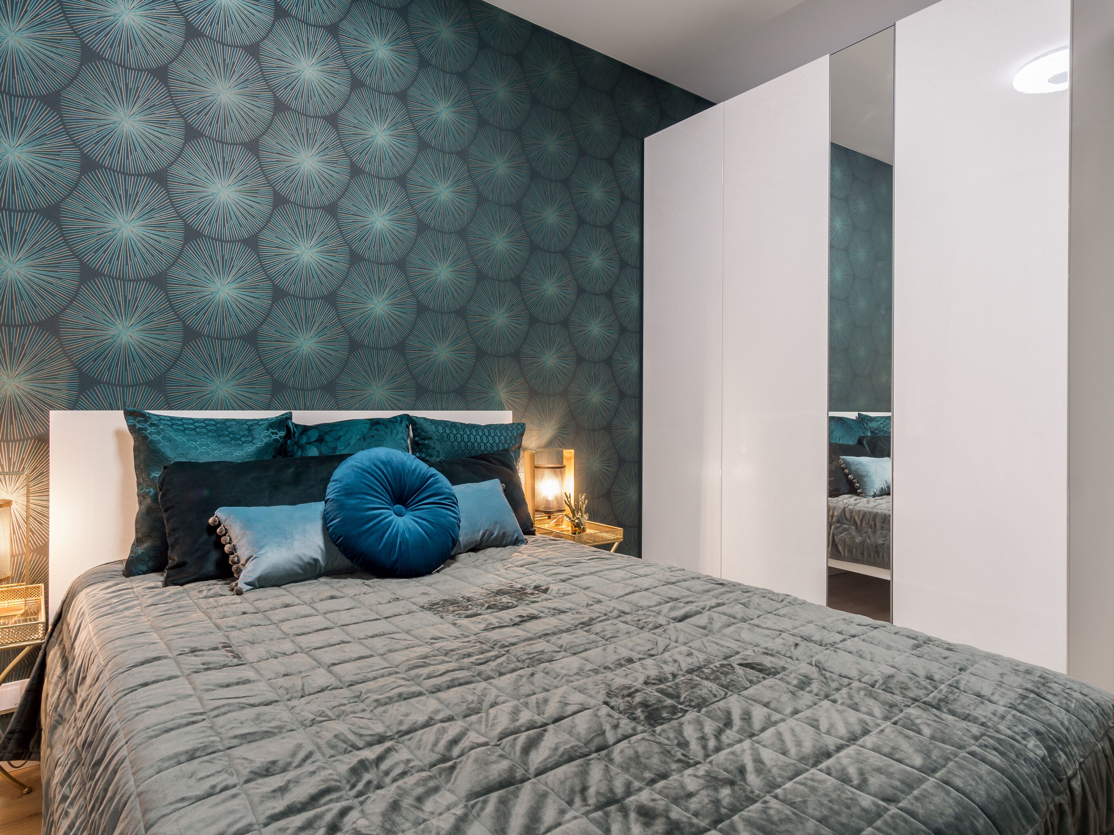 A modern bedroom with a patterned wallpaper, a large bed with multiple pillows, and a nightstand with a lamp.