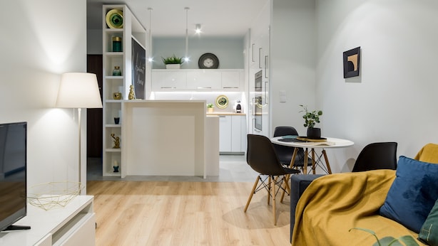 Una sala de estar moderna con televisor, sofá y mesa de centro. Se ve una cocina al fondo con armarios blancos y una zona de comedor.