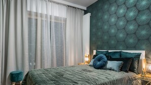 Un dormitorio con edredón verde acolchado, cabecero con almohadones decorativos, mesita de noche con lámpara y cortinas translúcidas en la ventana.
